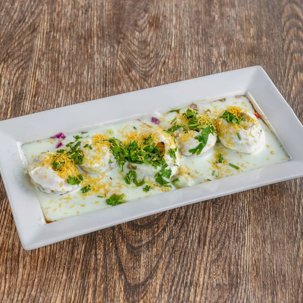 A white rectangular plate of Dahi Bhalla, topped with yogurt, cilantro, and sev, on a wooden table.