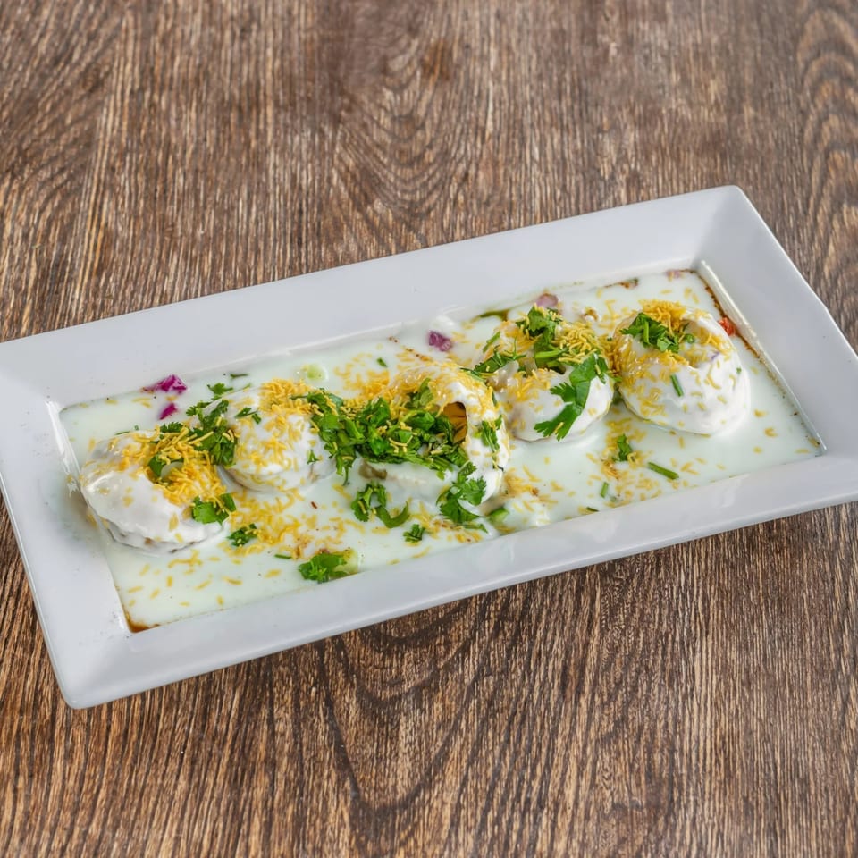 A white rectangular plate of Dahi Bhalla, topped with yogurt, cilantro, and sev, on a wooden table.